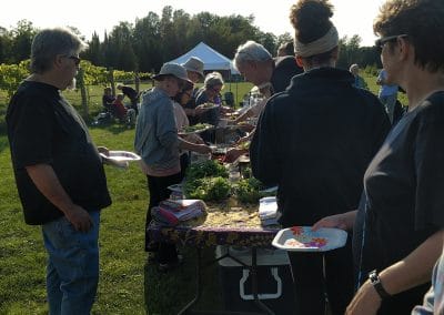 people gathered around a table in the vineyard, sharing a potluck meal