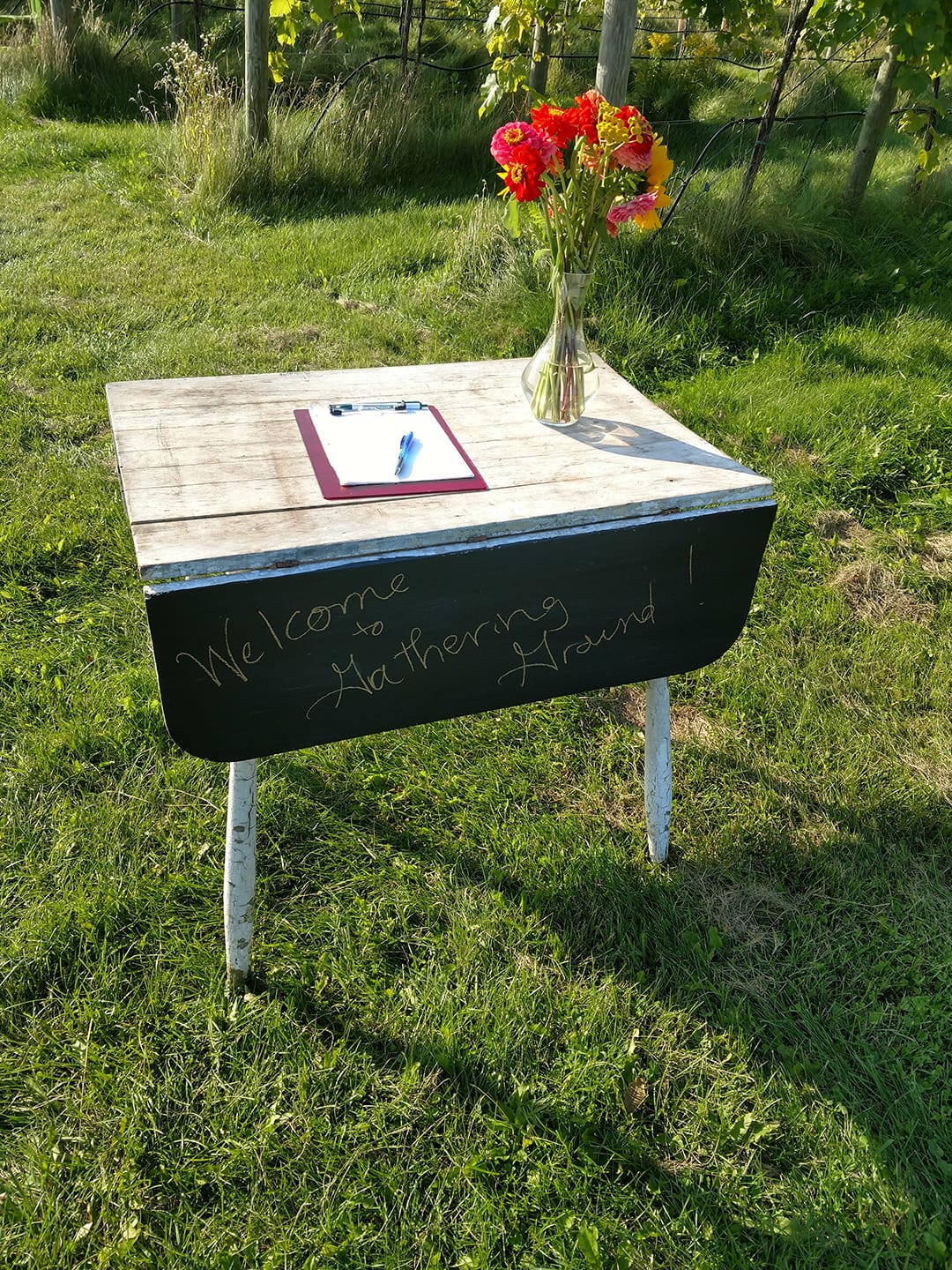 small table with "Welcome to Gathering Ground" written on it in chalk, a vase with dahlias, and a clipboard