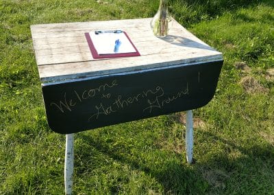 small table with "Welcome to Gathering Ground" written on it in chalk, a vase with dahlias, and a clipboard