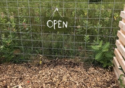 A compost pile is pictured in the foreground, with a sign reading "open" hanging from a fence, and an open field in the background.
