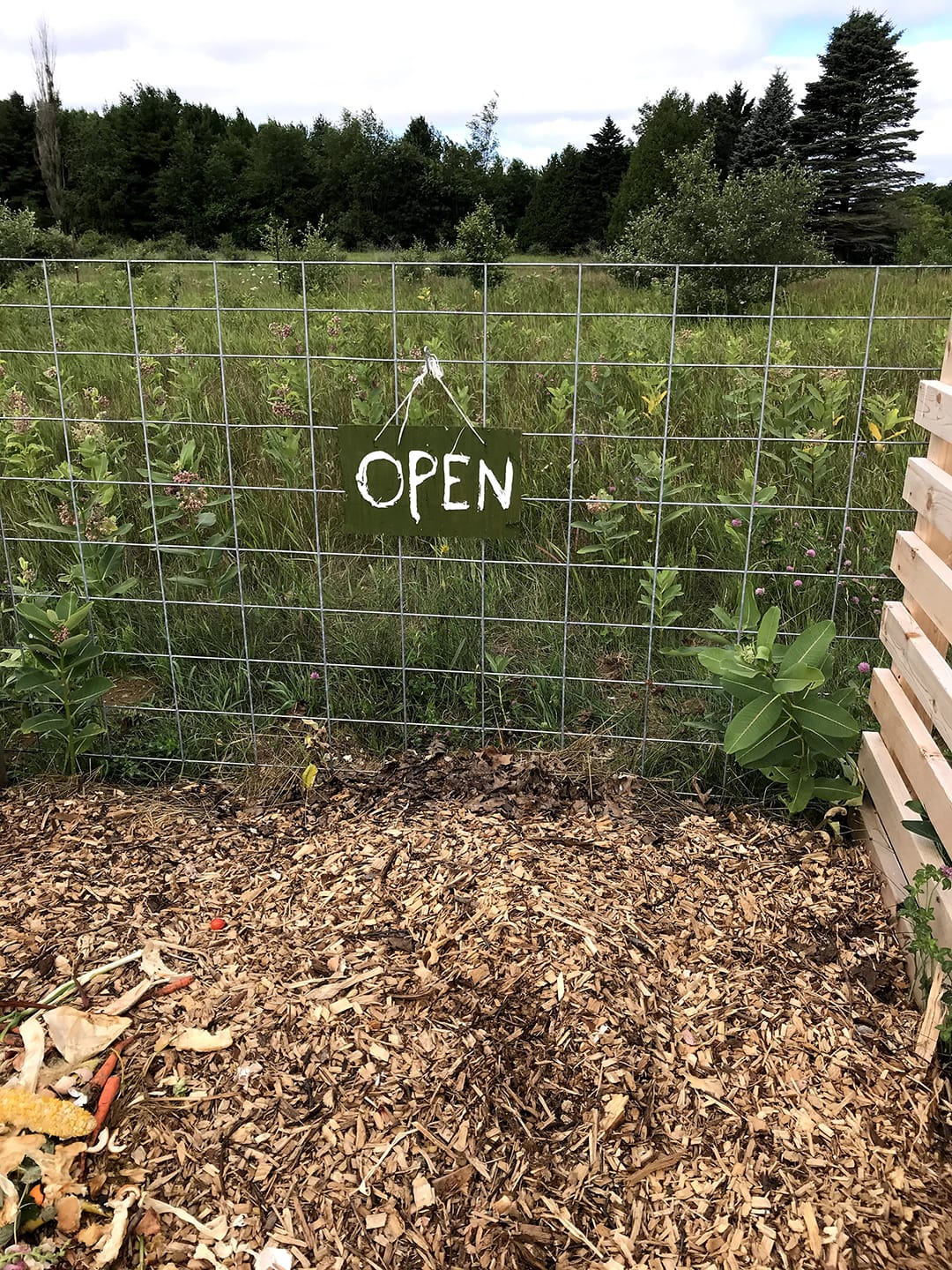 A compost pile is pictured in the foreground, with a sign reading "open" hanging from a fence, and an open field in the background.